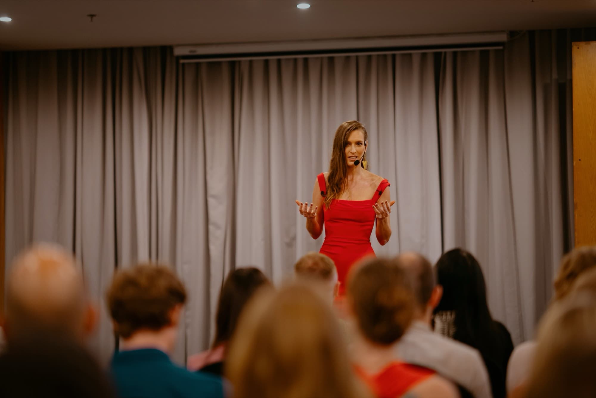 Wide view of a room with Arja Hendrikx in a red dress on a low stage and the audience seated along one side